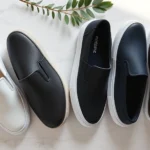Overhead flat lay of four premium slip-on sneakers in white, black, navy, and cream arranged on marble surface with minimalist styling elements, natural lighting