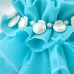 Close-up detail of Lagoona Blue doll's turquoise layered tail skirt fabric with iridescent shell embellishments and pearl-tone trim, showing miniature garment construction techniques and material quality, professional product photography style