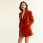 Woman wearing 1970s burnt orange velvet blazer with modern black ankle boots and minimal gold jewelry, standing against neutral backdrop, natural lighting, sophisticated vintage styling