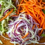 Overhead shot of freshly shredded green and red cabbage with julienned carrots on wooden cutting board, professional kitchen lighting, vibrant colors, shallow depth of field