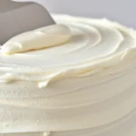 Close-up of creamy white powdered sugar frosting being spread smoothly onto a vanilla layer cake with an offset spatula, professional bakery lighting, detailed texture showing smooth frosting surface, neutral kitchen background