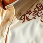 Close-up of premium western pearl snap shirt in cream with intricate embroidered yoke detail, rich texture visible on high-quality cotton fabric, warm studio lighting highlighting craftsmanship