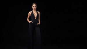 Woman wearing sleek black wide-leg flared jumpsuit with plunging neckline and gold layered jewelry, standing confidently in studio lighting, black background, professional fashion photography