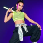 Model wearing neon lime green mesh crop top with black oversized cargo pants, chunky silver platform sneakers, holographic windbreaker tied at waist, multiple silver chain necklaces, glow sticks accessory, confident dancing pose, dark club lighting background with purple neon glow