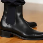 Close-up of black leather Chelsea boot showing elastic side panel and sleek silhouette, worn on polished hardwood floor, professional studio lighting highlighting leather texture and craftsmanship