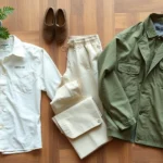 Flat lay of sustainable clothing items including organic cotton shirt, linen pants, and recycled fabric jacket arranged artfully on natural wood surface with green plants