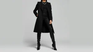 Person wearing all-black outfit with sharp architectural silhouette, dramatic long coat with structured shoulders, pointed-toe boots, and bold silver chain jewelry against neutral background. Professional fashion photography, high-contrast lighting.