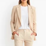 Woman in minimalist neutral outfit wearing tailored beige blazer, white t-shirt, and cream trousers, standing against white background, professional styling, natural lighting, clean aesthetic, fashion editorial style