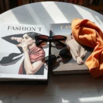 Flat lay of luxury fashion coffee table books with silk scarves and designer sunglasses arranged on marble surface, natural lighting, elegant aesthetic, high-end bookshelf styling
