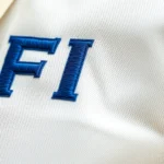 Close-up of a high-quality embroidered FIT logo on a cream-colored premium cotton shirt, showing precise stitching detail and texture, professional fashion photography style