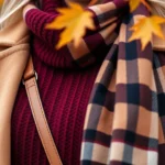 Close-up of rich burgundy wool sweater with camel coat layered over it, styled with cognac leather crossbody bag and oversized plaid scarf, autumn leaves in soft-focus background